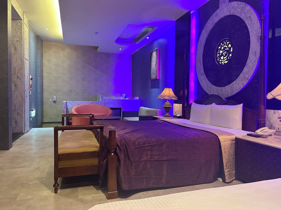 Una habitación de hotel moderna con una cama grande, un área de descanso con una silla y una mesita, un baño con espejo y lavamanos, y una pared decorativa con un diseño circular.