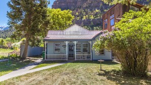 A small, single-story house with a porch, surrounded by trees and a mountain backdrop. - Cozy Downtown Pet Friendly Home with Fiber Internet! Fenced Outdoor Area! (Ouray)