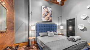 A bedroom with a blue headboard, a gray bedspread, a framed artwork above the bed, a round side table, a mirror with a wooden frame, and a door with a black handle.