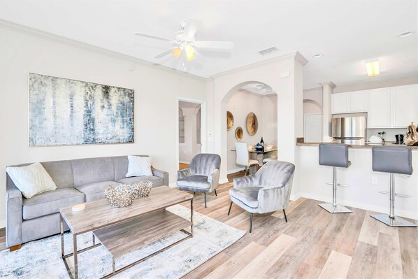 A modern living room with a grey sofa, a coffee table, and two grey armchairs. A kitchen area with white cabinets and a bar stool is visible in the background.
