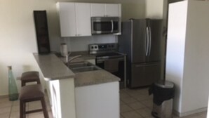 A compact kitchen with white cabinets, a microwave, a refrigerator, and a sink. There are two stools and a trash can. - PR-PARADISE, Serenity-3-BEDROOM-2 BathRM- APT -PARKING, POOL-WiFI-close to BEACH (Rio Grande)