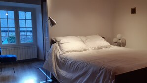 Een slaapkamer met een bed, een bureau en een stoel. Er is een raam met zonwering, een lamp en een wandlamp.