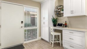A compact kitchen with white cabinetry, a dining area with a white chair, and a small table with a laptop and wine bottle. - Fashion Square Retreat | Walk Everywhere + 4 Pools (Scottsdale)