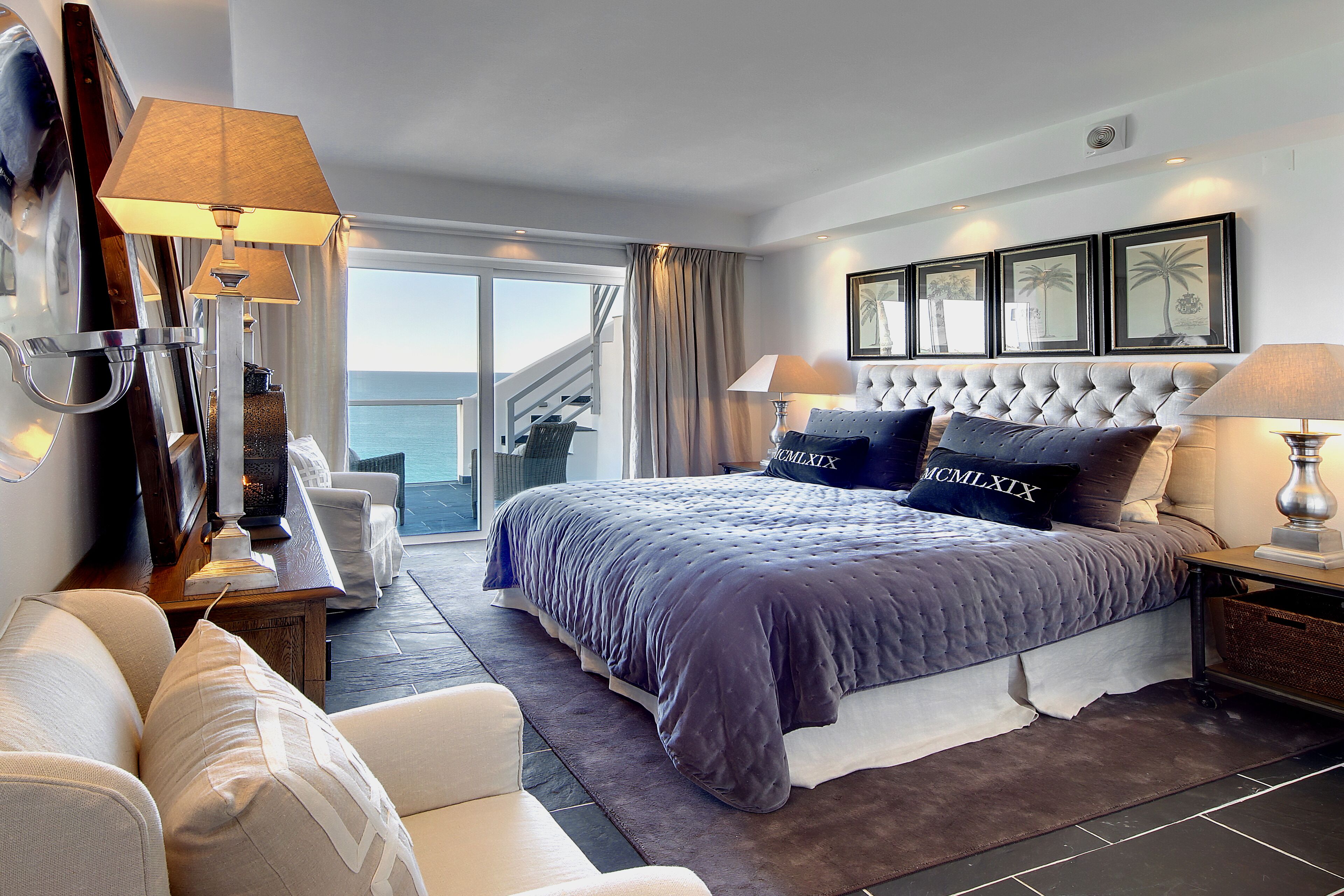 A modern hotel room with a large bed, a sitting area with a sofa and armchairs, a view of the sea through large windows, and a balcony.