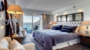 Una camera d'albergo moderna con un letto grande, una zona relax con divano e poltrone, vista mare attraverso ampie finestre e un balcone.