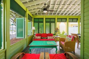 Un porche techado con una decoración verde y blanca, un sofá rojo con cojines, una mesa verde con un tablero de ajedrez, sillas de mimbre y un ventilador de techo.