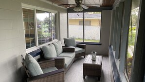 Eine überdachte Terrasse mit Korbmöbeln, einem Deckenventilator und großen Fenstern mit Blick ins Freie.