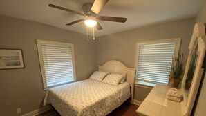 Ein Schlafzimmer mit einem Bett, Deckenventilator, Kommode und zwei Fenstern mit Jalousien.