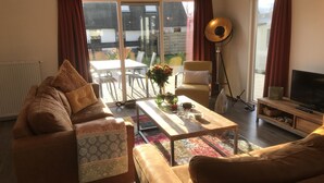 Ein Wohnzimmer mit einer Couch, einem Couchtisch und einem Fernseher. Eine Glasschiebetür führt zu einem Außenbereich mit Tisch und Stühlen.