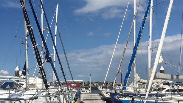 Ein Yachthafen mit mehreren angemeldeten Segelbooten, ein hölzerner Steg, der zum Wasser führt, und ein klarer, blauer Himmel.