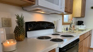 Une cuisine avec un four blanc, des placards en bois, un four à micro-ondes placé au-dessus du four, un ananas sur le comptoir et une bougie allumée.