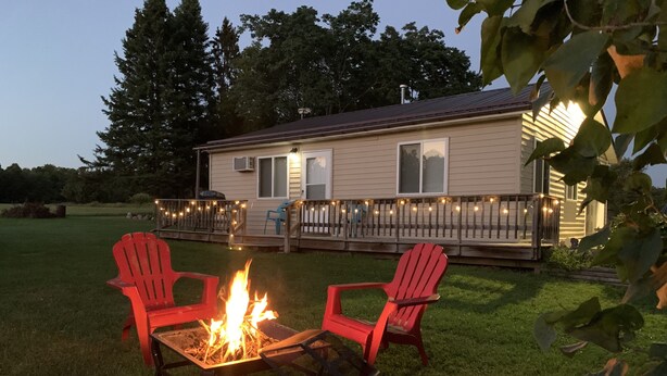 Un espace extérieur accueillant avec un foyer et deux fauteuils Adirondack rouges, une terrasse en bois illuminée de guirlandes lumineuses, et une maison en arrière-plan.