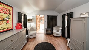 Quarto de hotel com um pôster do Mickey Mouse, duas poltronas, piso de madeira, tapete preto, guarda-roupa e um aviso indicando disponibilidade de Wi-Fi.