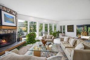 Un salon spacieux avec une cheminée en pierre, de grandes fenêtres, une table basse en verre et un canapé beige.