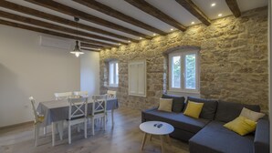 Une pièce avec un mur en pierre, des poutres en bois, un canapé gris agrémenté de coussins jaunes, une table blanche avec des chaises et une fenêtre avec des volets blancs.