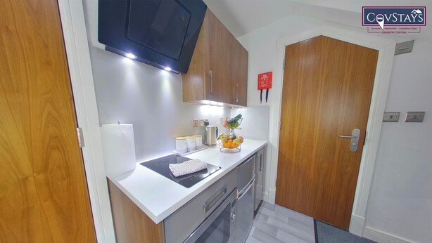 A compact kitchen with a built-in oven, a microwave, and a sink. There is a small countertop with a kettle and a fruit bowl. The kitchen has wooden cabinets and a white countertop. - Victoria House - Deluxe Studio by CovStays (Coventry)