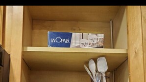 A wooden cabinet with three shelves. The top shelf has a box labeled "La Opala." The middle shelf has a box labeled "simply done" and a set of kitchen utensils. The bottom shelf has a bottle of oil, spice jars, and a container of flour. - Townhome in gated community nearby a lot of attraction shopping and restaurants  (Sandy Springs)