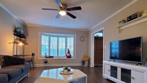 A modern living room with a ceiling fan, a large window with blinds, a sofa, a coffee table, a TV on a white stand, and a small shelf with decorative items. - Townhome in gated community nearby a lot of attraction shopping and restaurants  (Sandy Springs)