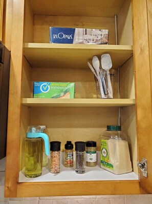 A wooden cabinet with three shelves. The top shelf has a box labeled "La Opala." The middle shelf has a box labeled "simply done" and a set of kitchen utensils. The bottom shelf has a bottle of oil, spice jars, and a container of flour. - Townhome in gated community nearby a lot of attraction shopping and restaurants  (Sandy Springs)