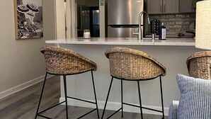 Una cocina moderna con un refrigerador de acero inoxidable, horno empotrado y microondas. Hay dos taburetes de mimbre y un sofá a rayas.