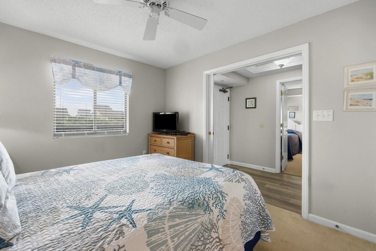 Une chambre à coucher avec un lit, un téléviseur posé sur une commode en bois, un ventilateur de plafond et une porte donnant sur une autre pièce.