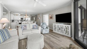 Ein modernes Wohnzimmer mit einem grauen Sofa, einem Hocker und einem gemusterten Sessel. Ein Flachbildfernseher ist an einem weißen Schrank befestigt, und ein Deckenventilator ist sichtbar.