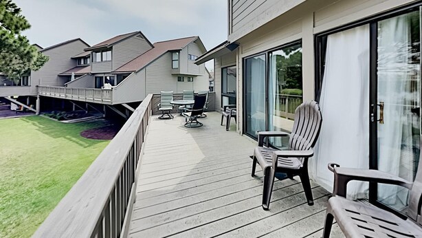 Una terraza de madera con mobiliario exterior, una puerta de vidrio que da a un espacio cubierto y vistas a casas vecinas.