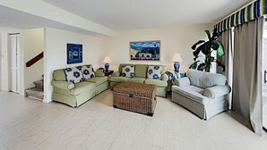 Una sala de estar con un sofá modular, un sillón, un reposapiés de mimbre y una planta grande en maceta.