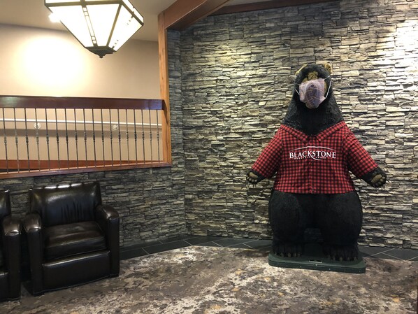 A plush bear statue wearing a red and black checkered shirt with the text "BLACKSTONE MOUNTAIN LODGE" in a lobby area with stone walls and a wooden railing.