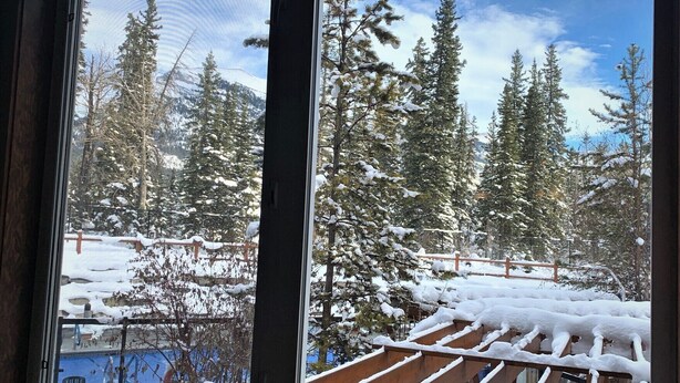A view through a window of a snowy landscape with trees and a wooden deck. - Spacious (700 sqft) 1 Bedroom/1 Bath cozy Condo /Pool & Hot tubs (Canmore)