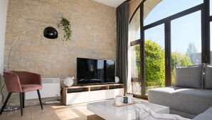 Una sala de estar moderna con una pared de piedra, un televisor de pantalla plana, un sillón rosa, un sofá gris y ventanas amplias con vistas a la vegetación.