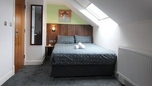 A bedroom with a sloped ceiling, a bed with patterned bedding, a nightstand, a wall-mounted lamp, and a painting.