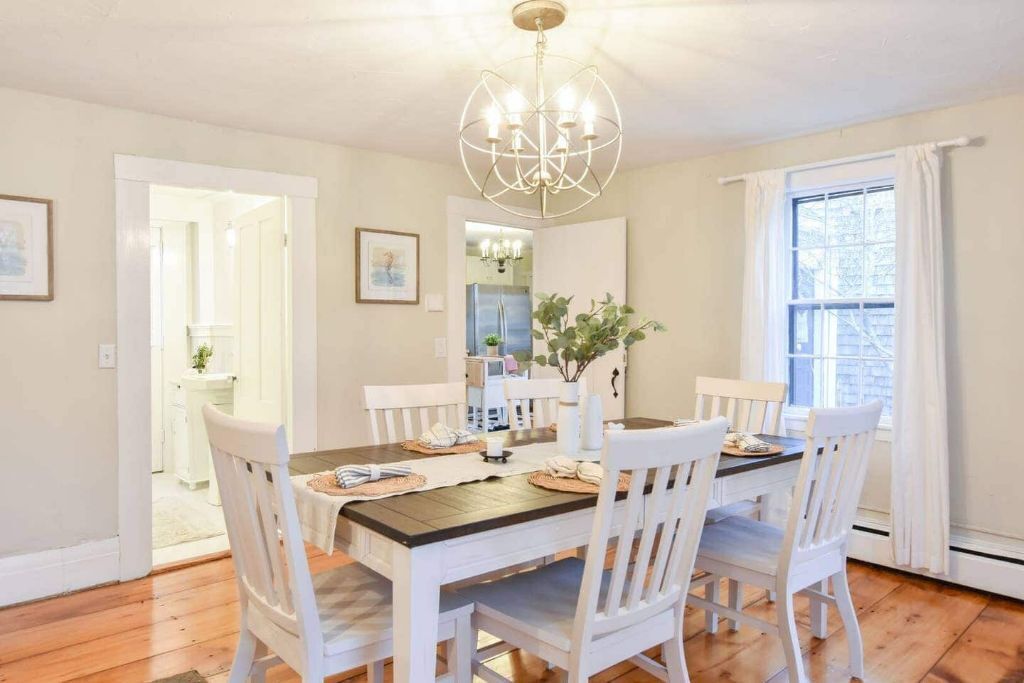A dining area with a wooden table, white chairs, a chandelier, and a window with curtains.