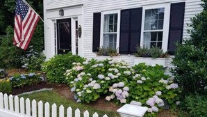 Une maison blanche avec des volets noirs, une clôture en piquets blancs et des arbustes d’hortensias.