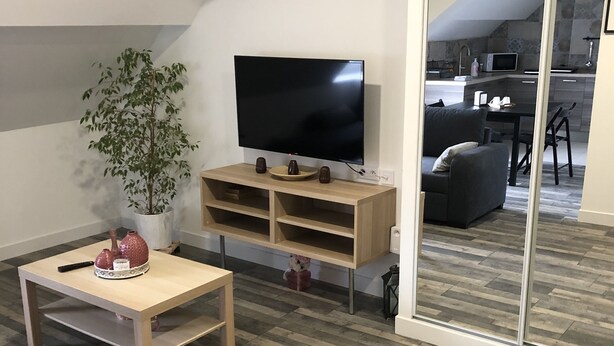 A modern living room with a flat-screen TV, wooden furniture, a potted plant, and a view into the kitchen.