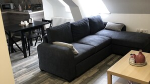 A modern loft apartment with a dark grey sectional sofa, a wooden dining table, and a kitchen in the background.