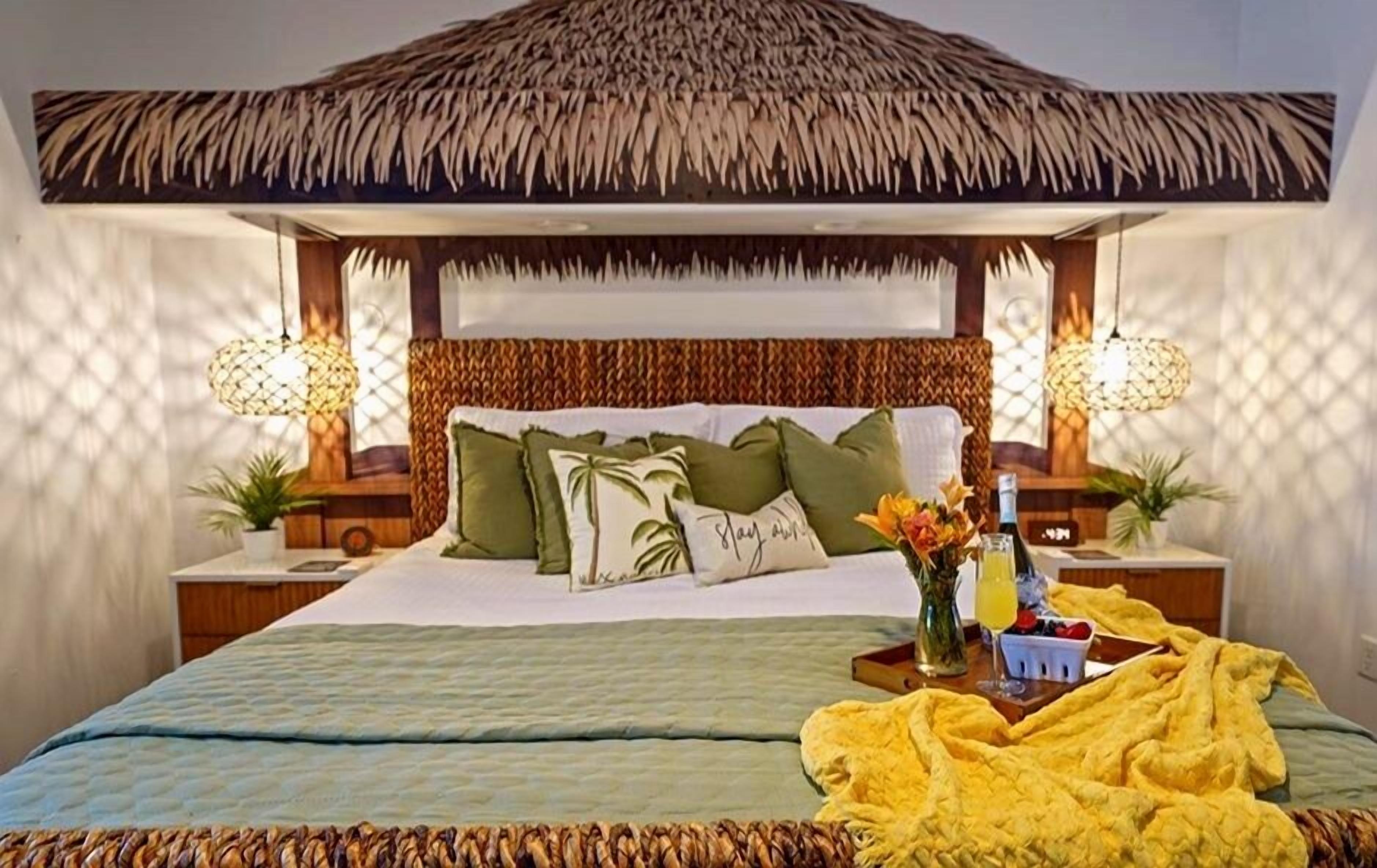 A bed with a thatched roof, two bedside lamps, and a tray with a drink and fruit.