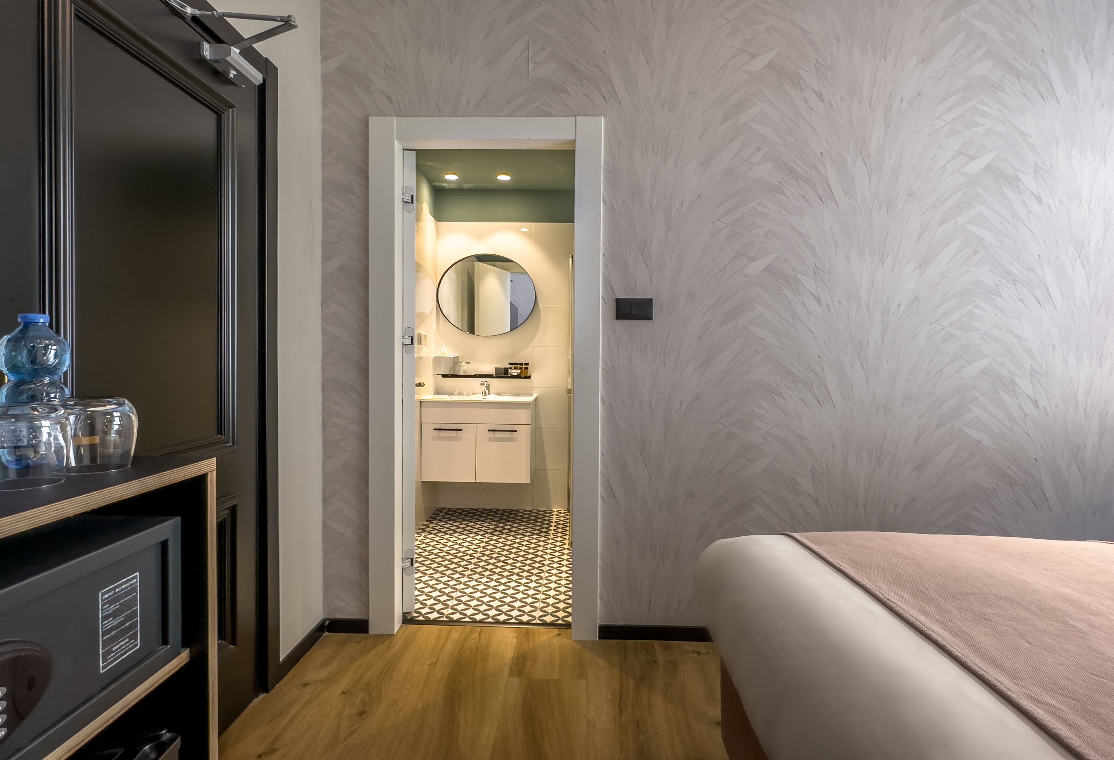 Una camera d'albergo con pavimento in legno, un letto con coperta beige, un mobile TV nero e un bagno con specchio rotondo e mobili bianchi.