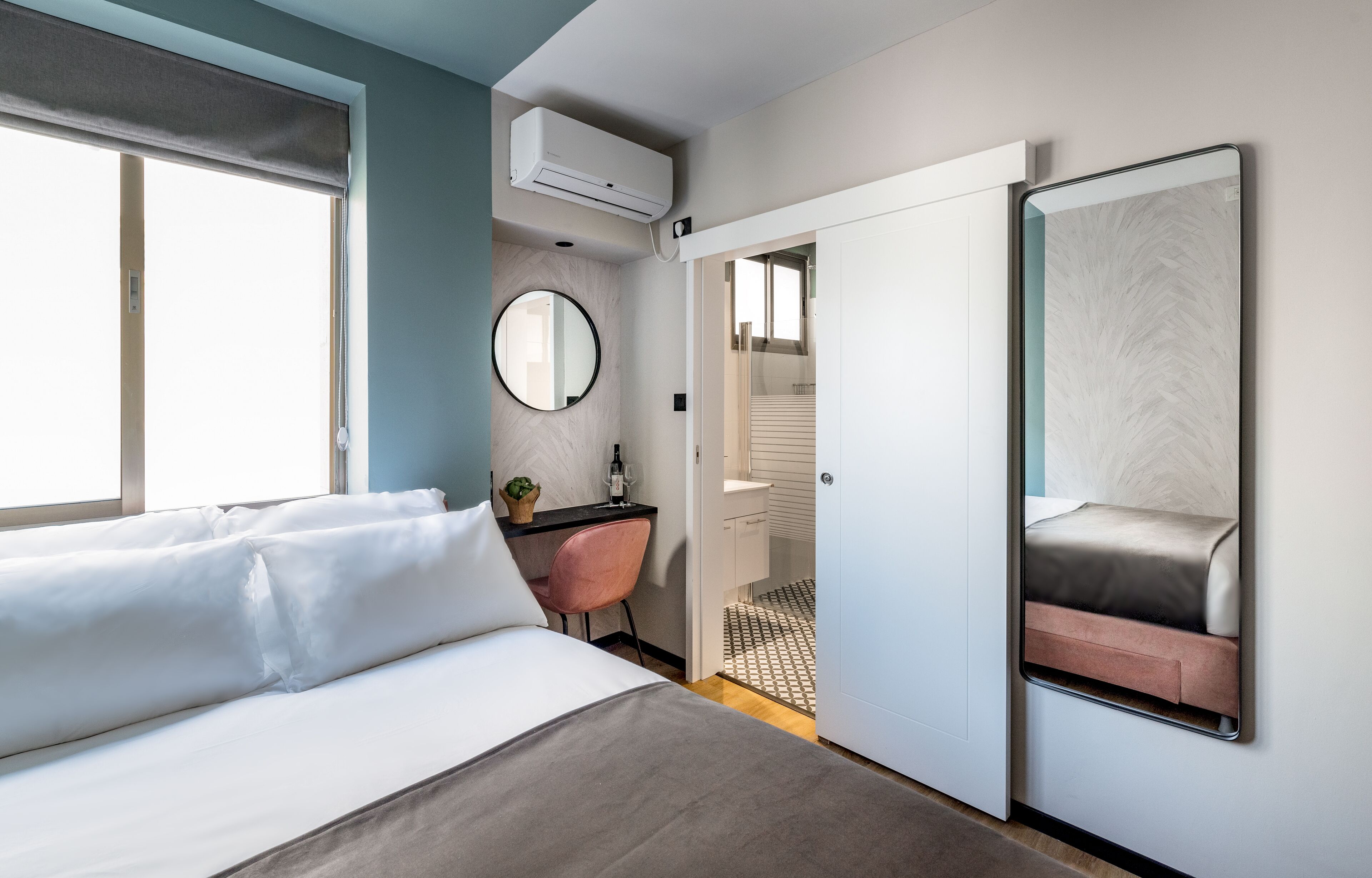 Moderni hotellihuone, jossa on sänky, työpöytä, peili ja ilmastointilaite.