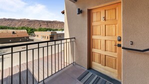 Une porte en bois avec une plaque portant un numéro, un balcon avec une rambarde en métal et une vue sur des bâtiments et une montagne en arrière-plan.