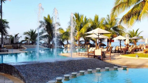 Une piscine dotée de jets d’eau, entourée de chaises longues et de parasols, avec des palmiers en arrière-plan.