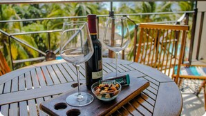 Une table en bois, avec deux verres à vin, une bouteille de vin et un bol de noix, sur un balcon offrant une vue sur des palmiers.