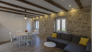 A spacious living area with a stone wall, a wooden ceiling, a gray sofa with yellow cushions, a white round table with chairs, and a window providing natural light. - Apartments Bepi-Apartment Roko (Komiža)