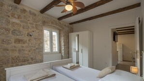 A bedroom with two beds, stone walls, wooden beams, a ceiling fan, and a window. - Apartments Bepi-Apartment Roko (Komiža)