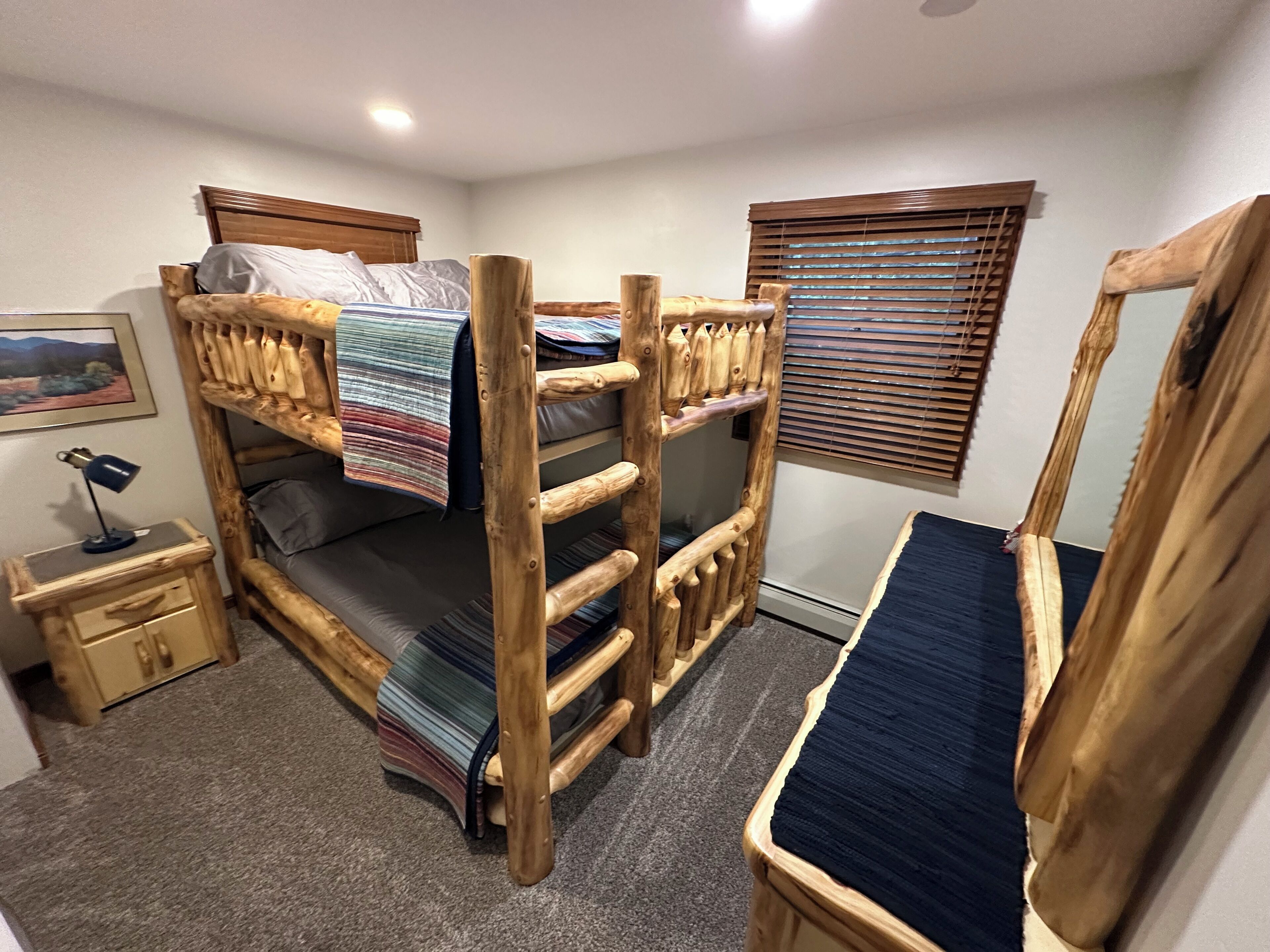 Habitación con literas, una escalera de madera, mesita de noche con lámpara y ventana con persianas.
