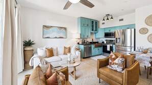 Un salon moderne avec un canapé blanc, des fauteuils en cuir marron et un coin repas comprenant une table et des chaises. On aperçoit, au fond, une cuisine avec des armoires de couleur turquoise et des appareils électroménagers en acier inoxydable.