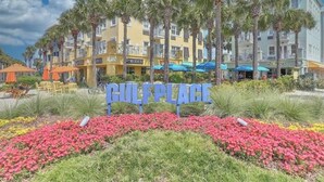 Un hôtel appelé « GULF PLACE », avec une enseigne devant l’entrée, entouré de palmiers et de parterres de fleurs aux couleurs vives.