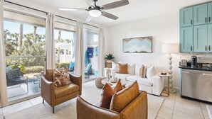 Un vaste espace de vie comprenant un canapé, des fauteuils et une table à manger. On y trouve également un ventilateur de plafond, une cuisine équipée de placards et une grande fenêtre avec des rideaux.