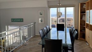 A dining area with a glass table and chairs, a view of the beach, and a sign reading "BEACH HOUSE". - Oceanfront 3BR/3BA with direct ocean views, private parking and bikes included (Cape May)