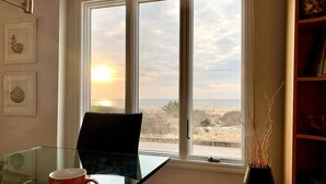 A room with a large window overlooking a beach, a glass table with a mug, and a bookshelf. - Oceanfront 3BR/3BA with direct ocean views, private parking and bikes included (Cape May)